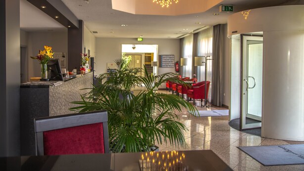 Um hall de hotel com balcão de recepção, cadeiras vermelhas, uma grande planta em vaso e um lustre moderno.