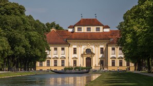 Um edifício grande e histórico, com fachada simétrica, entrada central com arco ornamentado e reflexo na água em frente.