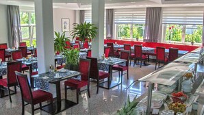 Um restaurante com cadeiras e mesas vermelhas, um buffet com frutas e bebidas, e grandes janelas com cortinas.