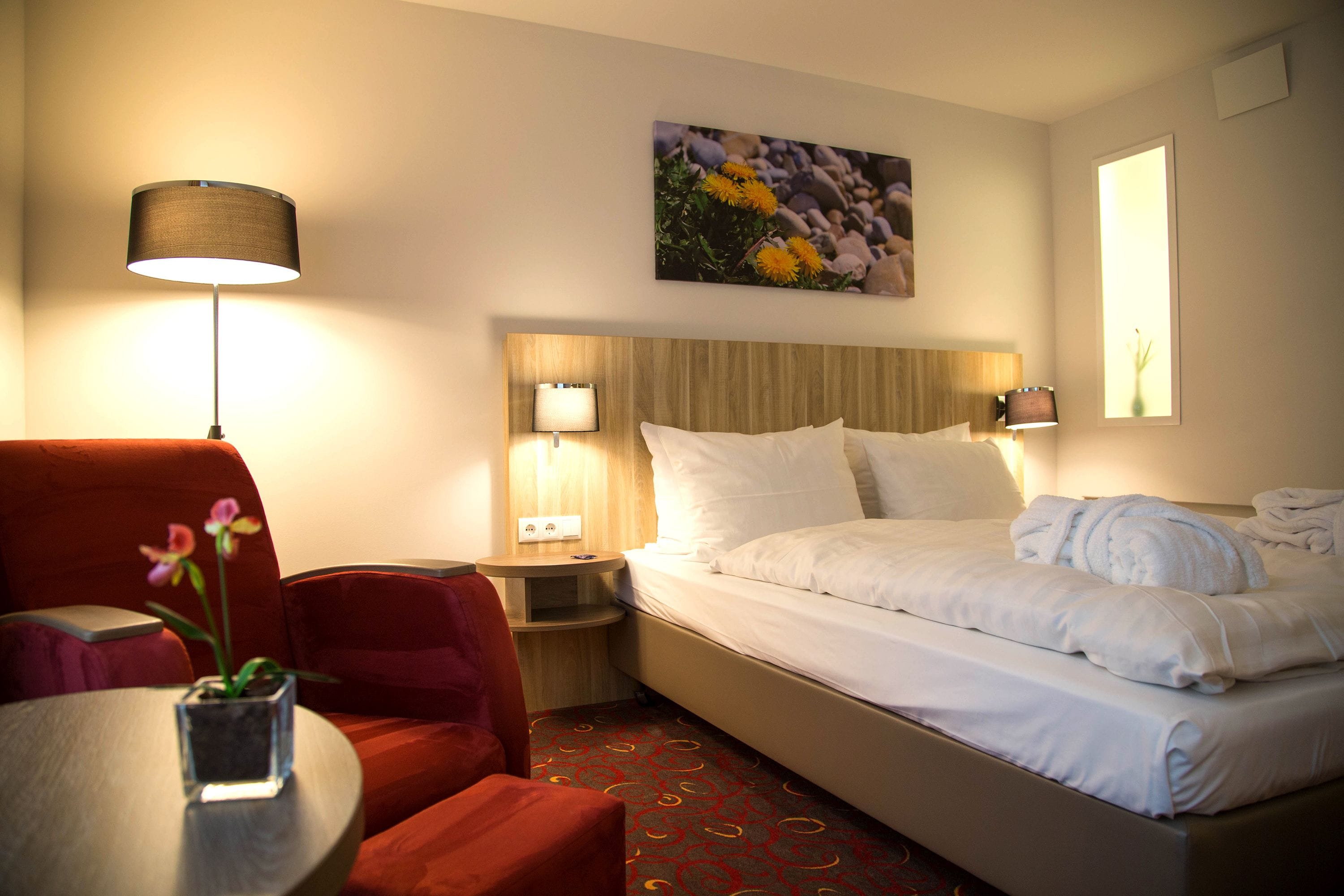 Una habitación de hotel con una cama, un escritorio, una silla, una mesa con un florero y un cuadro en la pared.
