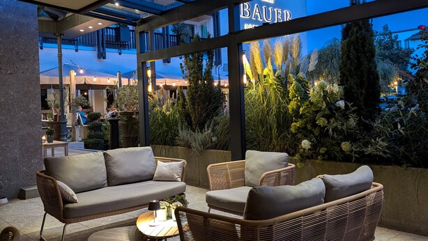 Un área de estar al aire libre con mobiliario de mimbre y techo de vidrio, con vistas a un jardín y un edificio con la señalización 'BAUER'.