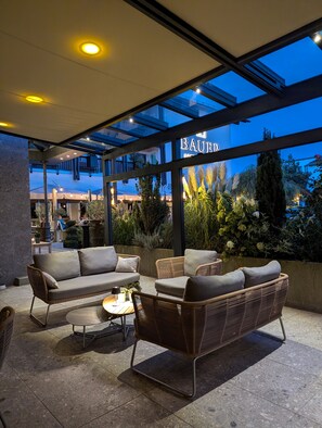 Un área de estar al aire libre con mobiliario de mimbre y techo de vidrio, con vistas a un jardín y un edificio con la señalización 'BAUER'.