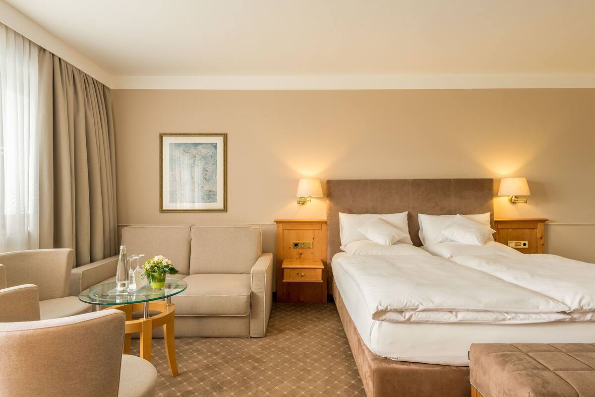 A hotel room with a bed, a sofa, a glass table with a vase, and two bedside lamps.