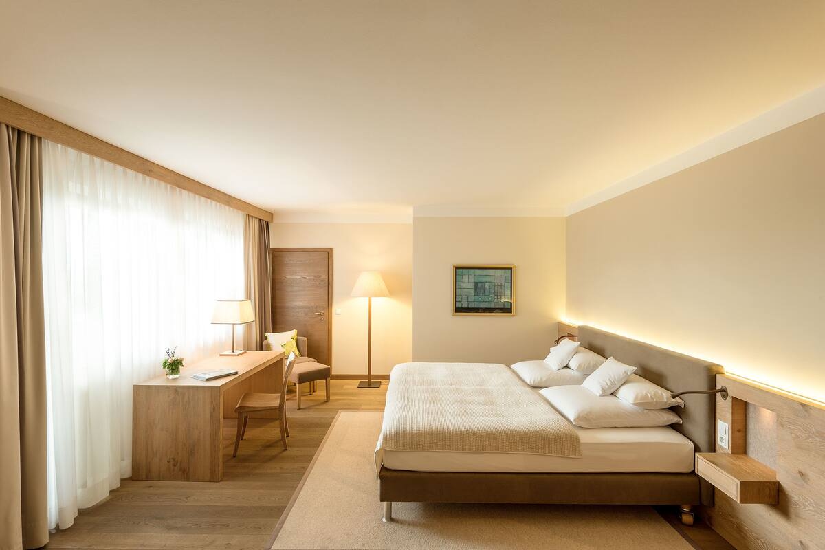 A hotel room with a bed, desk, chair, lamp, and a painting on the wall.
