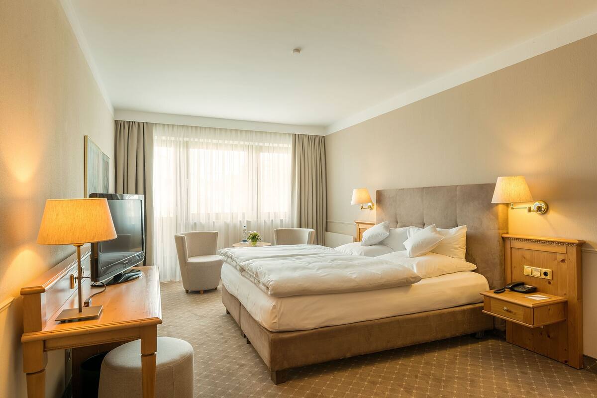 A hotel room with a large bed, a desk with a lamp, a chair, a TV, and a bedside table with a lamp and phone.