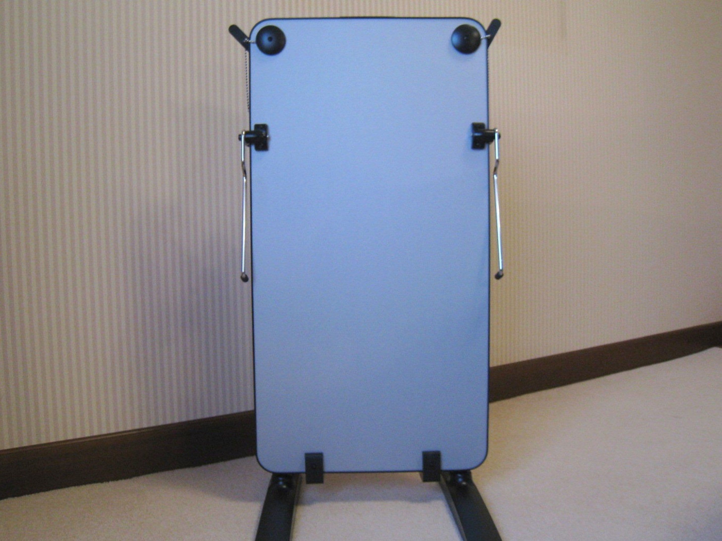 A whiteboard stand with two adjustable arms and a flat surface, mounted on a carpeted floor against a wall with vertical blinds.