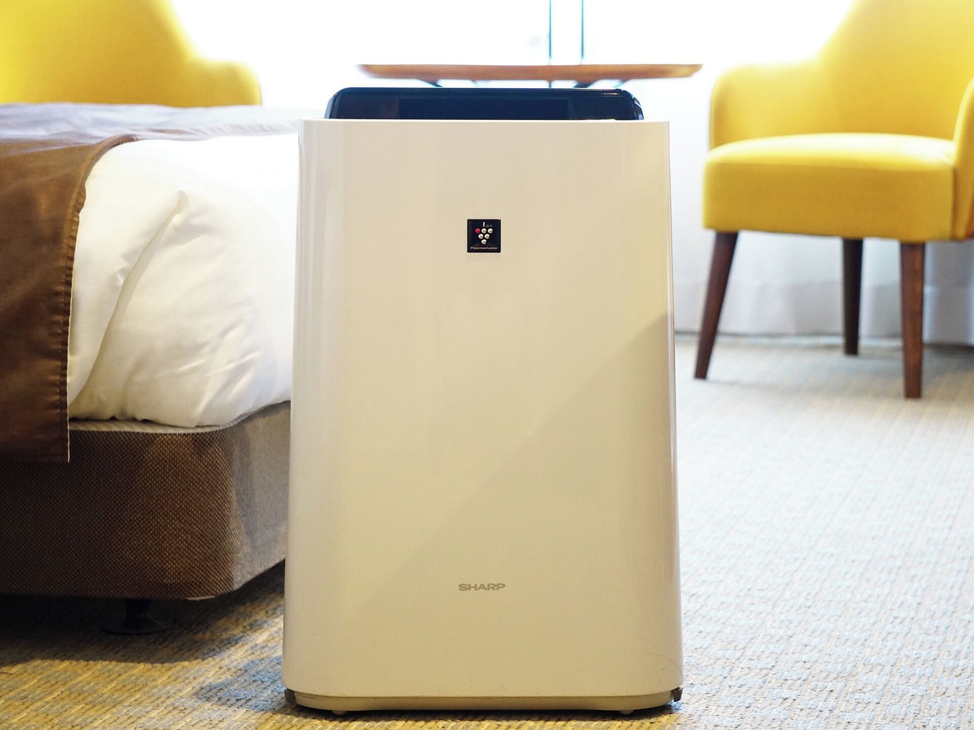 A white portable air purifier with a control panel on top, placed on a carpeted floor next to a bed with a brown headboard and white pillow.