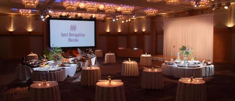 A banquet hall with round tables, chandeliers, and a projection screen displaying "Hotel Metropolitan Morioka".