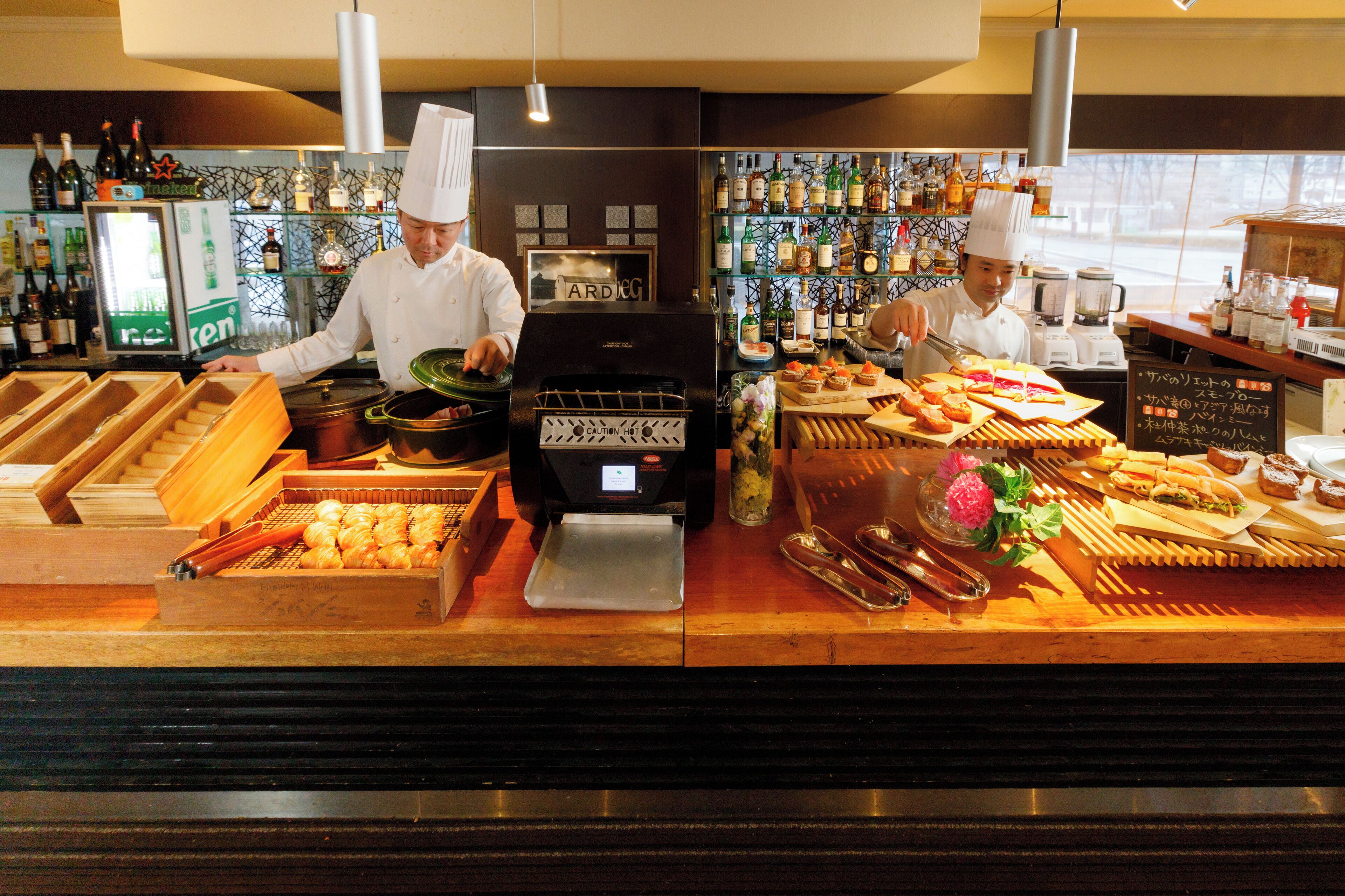 A kitchen counter with chefs preparing food, a refrigerator, and various cooking utensils.