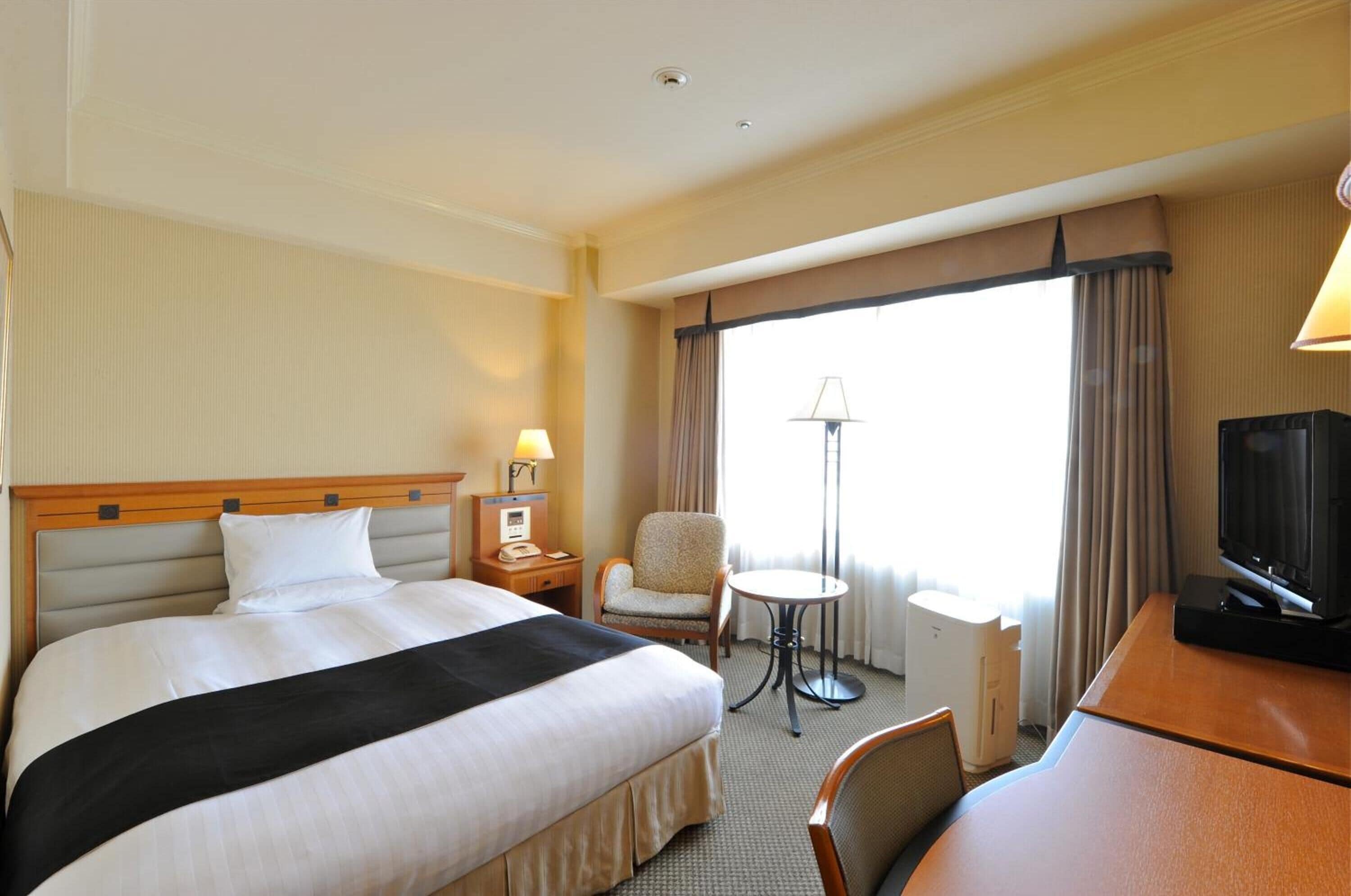 Habitación de hotel con una cama grande, un escritorio con televisor, una silla, una lámpara y un ventanal con cortinas.