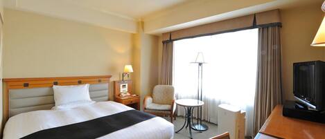 Habitación de hotel con una cama grande, un escritorio con televisor, una silla, una lámpara y un ventanal con cortinas.