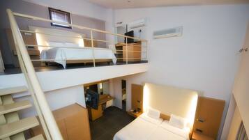 Une chambre à deux niveaux, avec un lit superposé à l’étage supérieur et un lit simple à l’étage inférieur. On y trouve un bureau avec une chaise, une armoire et une petite table avec une lampe.