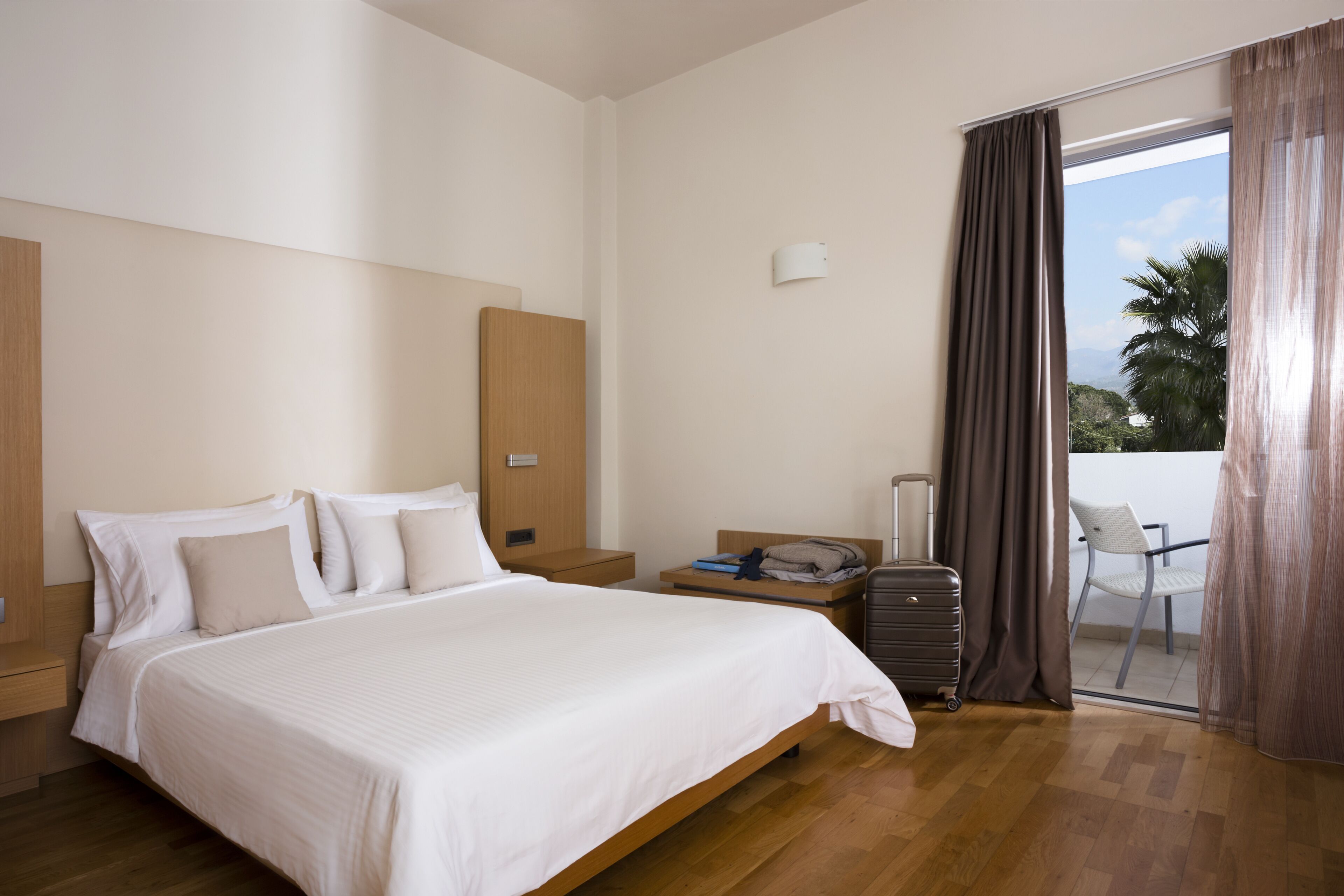 A hotel room with a large bed, a wooden headboard, a nightstand, a suitcase, and a view of the outdoors through a sliding glass door.