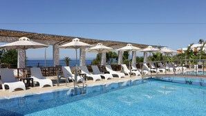 Un espace aménagé au bord de la piscine, avec des chaises longues blanches et des parasols, une piscine à l’eau d’un bleu limpide et une vue sur la mer.