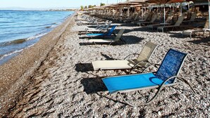 Une plage bordée de rangées de transats et de parasols, une plage de galets, et un bâtiment avec des balcons en arrière-plan.