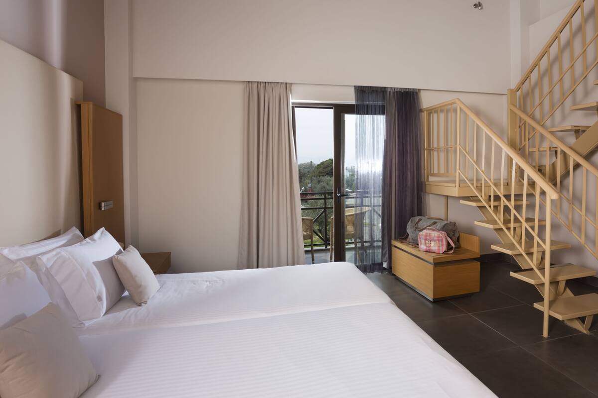 Habitación de hotel con una cama grande, un banco de madera, una escalera y un balcón con vistas.