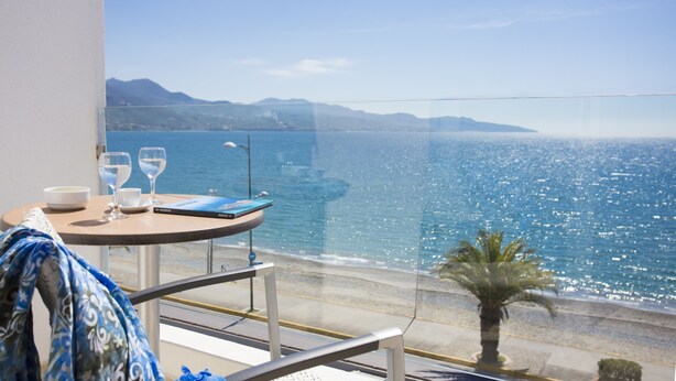 Un balcon avec une table dressée pour deux personnes, offrant une vue sur une mer d’un bleu limpide et sur des montagnes lointaines.