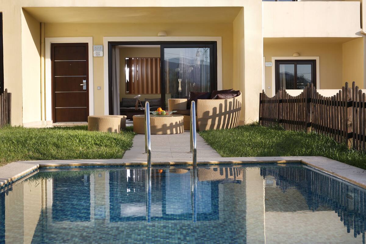 Una piscina con barrera de cristal, valla de madera y acceso a un edificio con puerta y ventana.
