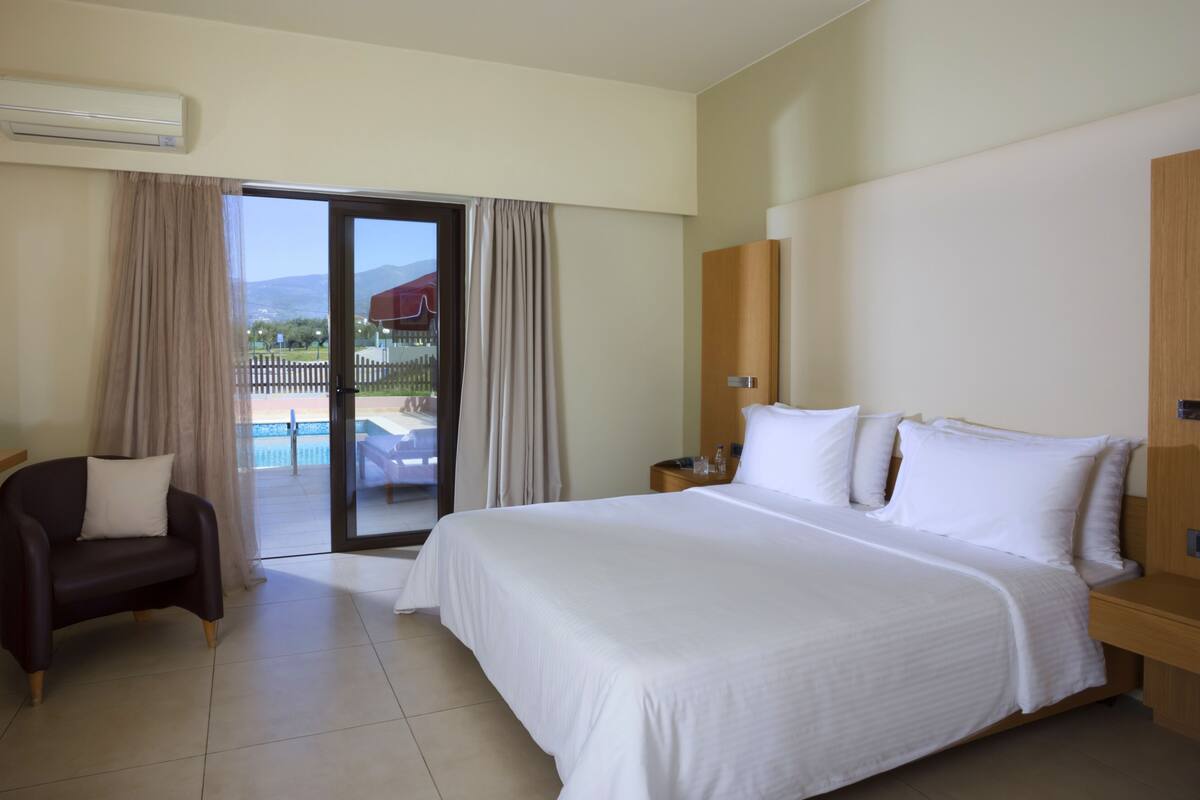 Habitación de hotel con una cama grande, una silla, una mesita de noche y vistas a una piscina y montañas.