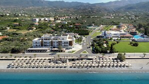 Vue aérienne d’une station balnéaire comprenant plusieurs bâtiments, une plage avec des transats et une piscine.