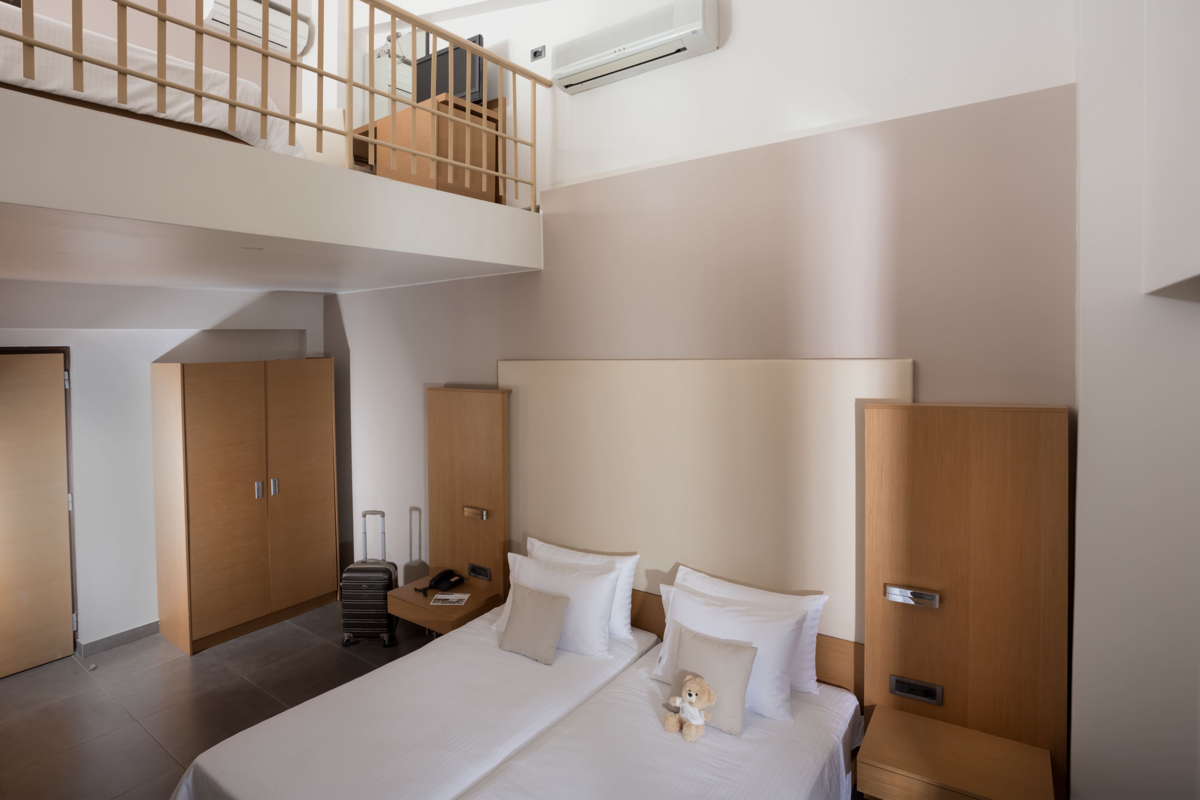 Habitación de hotel con literas, un escritorio, una silla, una maleta y un osito de peluche en la cama.