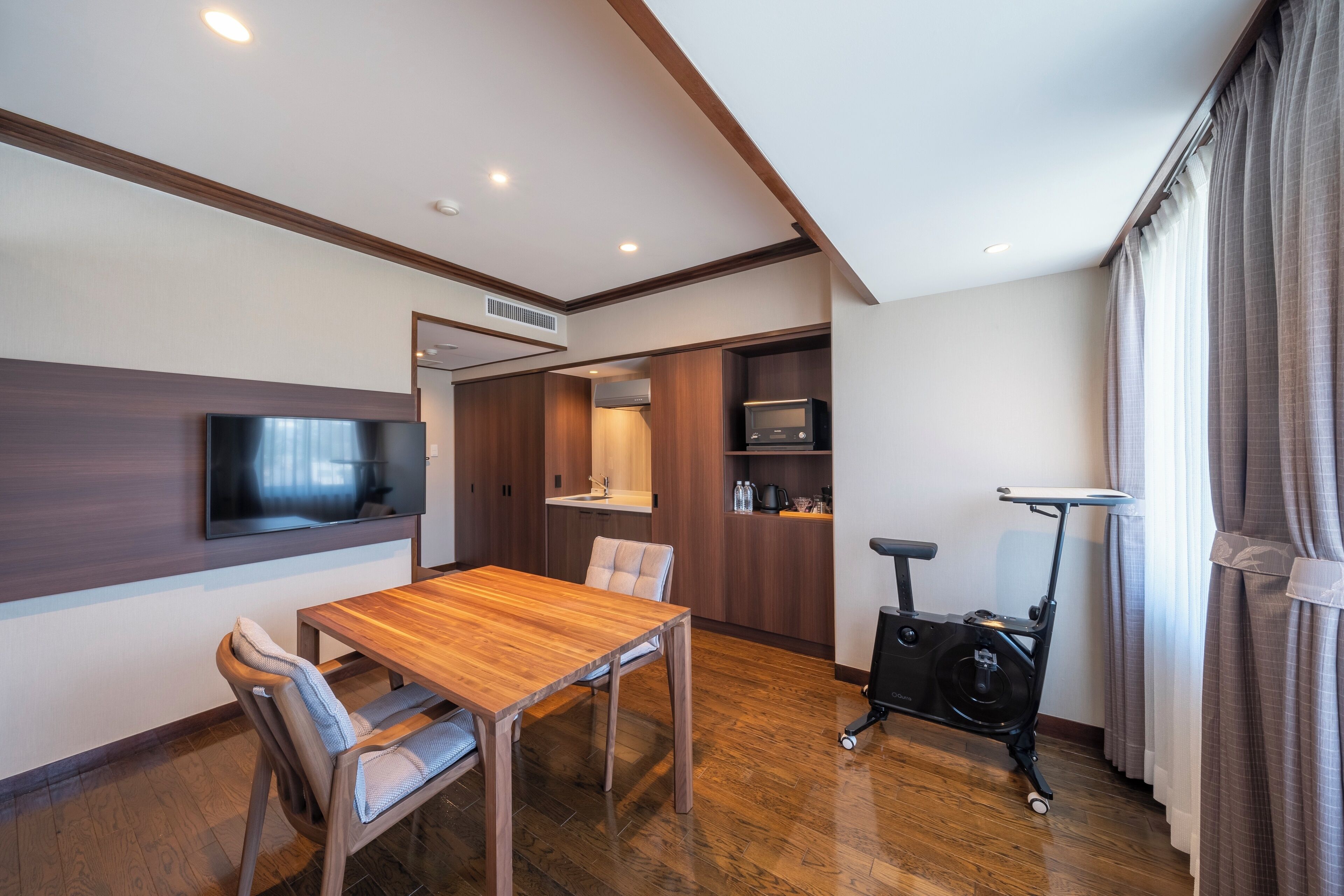 A modern living room with a wooden dining table, a flat-screen TV mounted on the wall, a built-in air conditioning unit, and a stationary exercise bike.