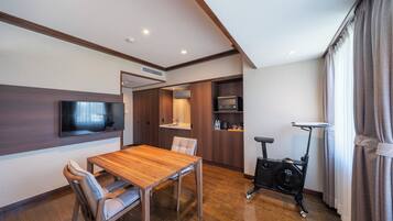 A modern living room with a wooden dining table, a flat-screen TV mounted on the wall, a built-in air conditioning unit, and a stationary exercise bike.