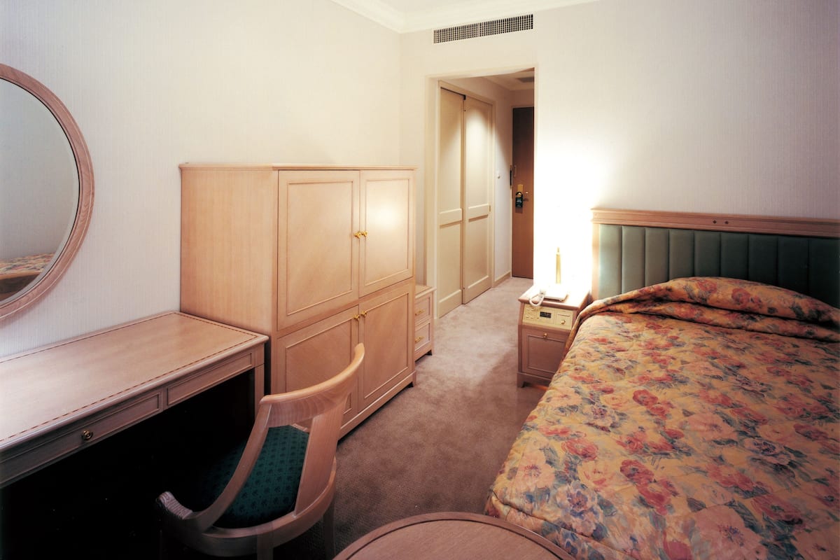 A hotel room with a wooden desk, a chair, a bed with floral bedding, a nightstand with a lamp, and a wardrobe.