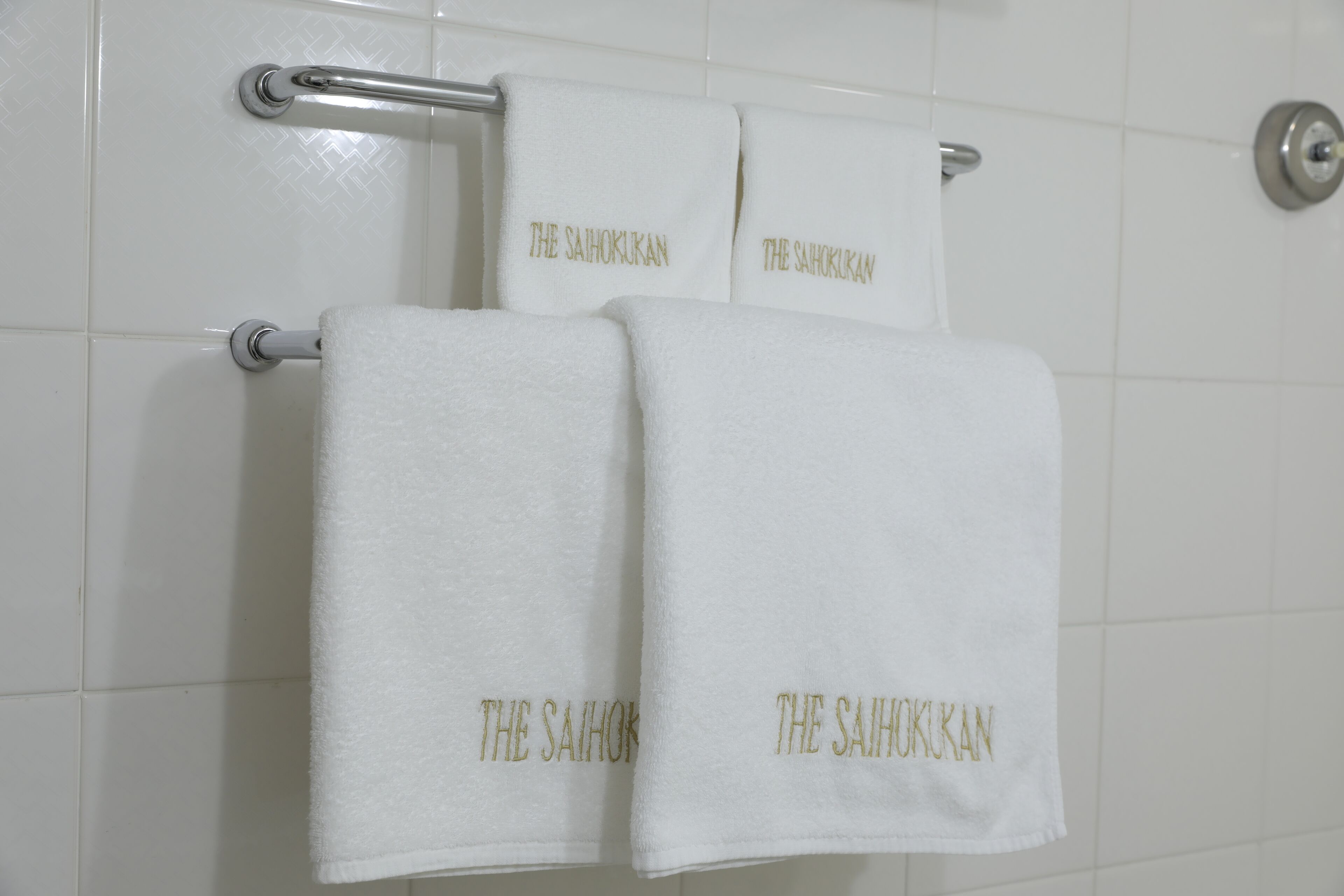 Two white towels with "THE SAIHOKUKAN" embroidered in gold, hanging on a metal towel rack in a bathroom with white tiled walls.