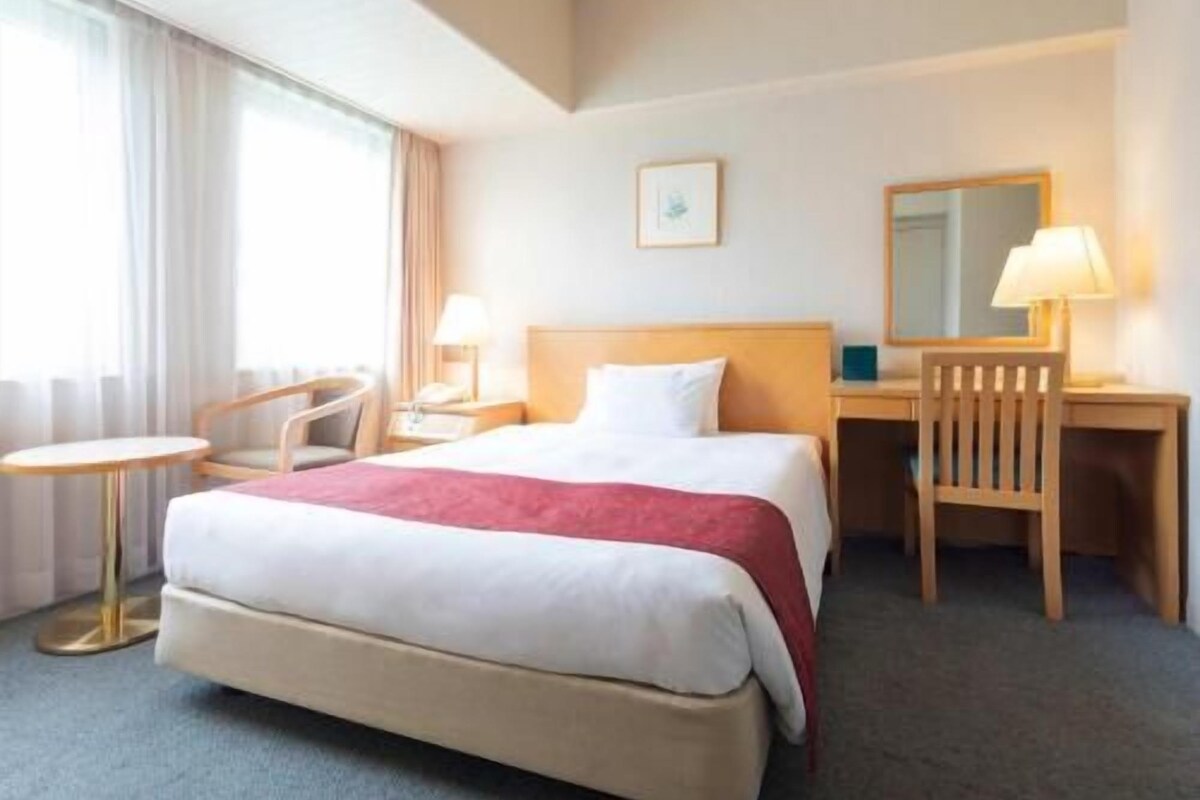 A hotel room with a bed, a desk, a chair, a small table, and a window with curtains.