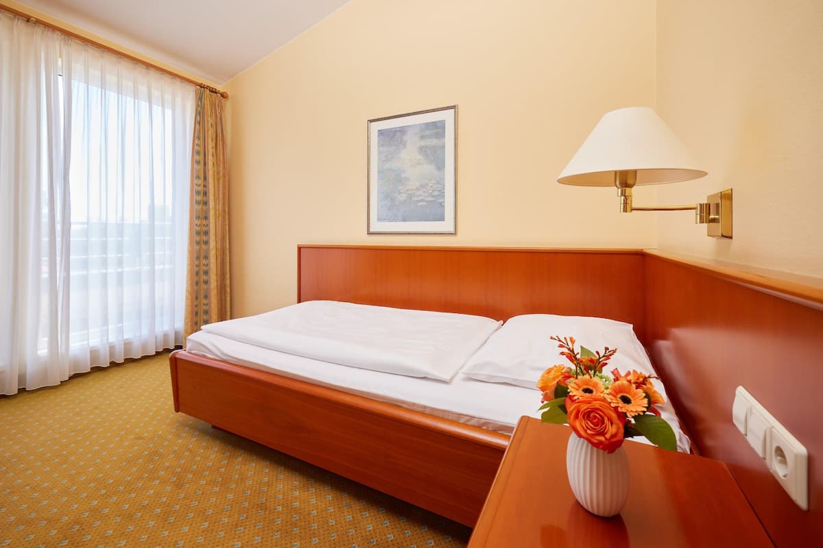 A hotel room with a wooden bed, a bedside table with a vase of flowers, a framed picture on the wall, and a window with sheer curtains.