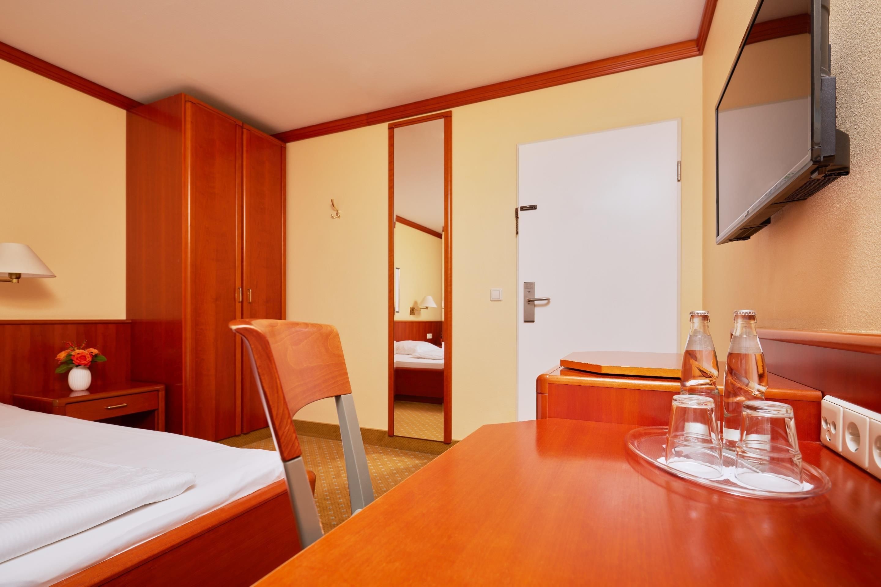 A hotel room with a bed, a desk, a chair, a wardrobe, a television, and a mirror.