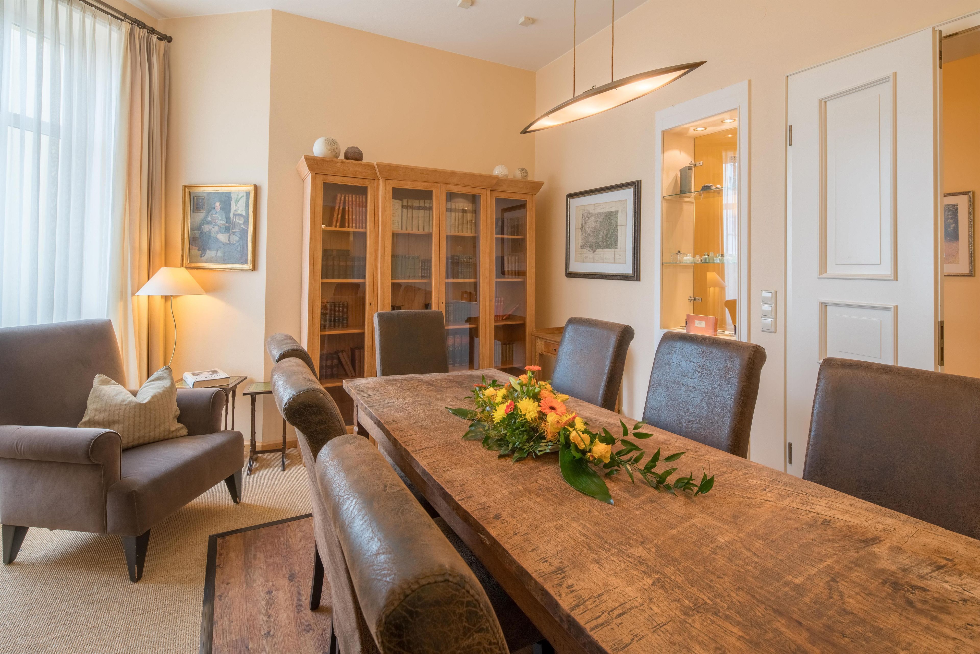 A dining area with a wooden table, chairs, a vase of flowers, and a bookshelf.