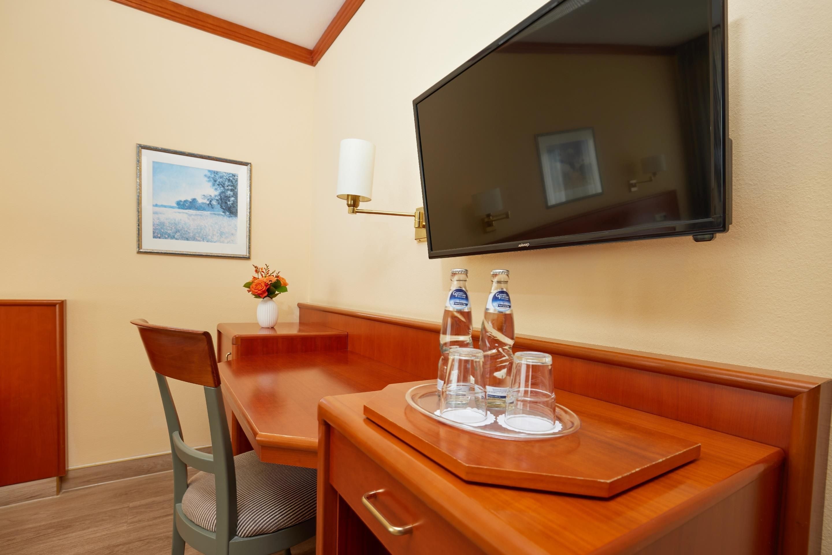 A hotel room with a wooden desk, a chair, a television mounted on the wall, and a framed picture on the wall.