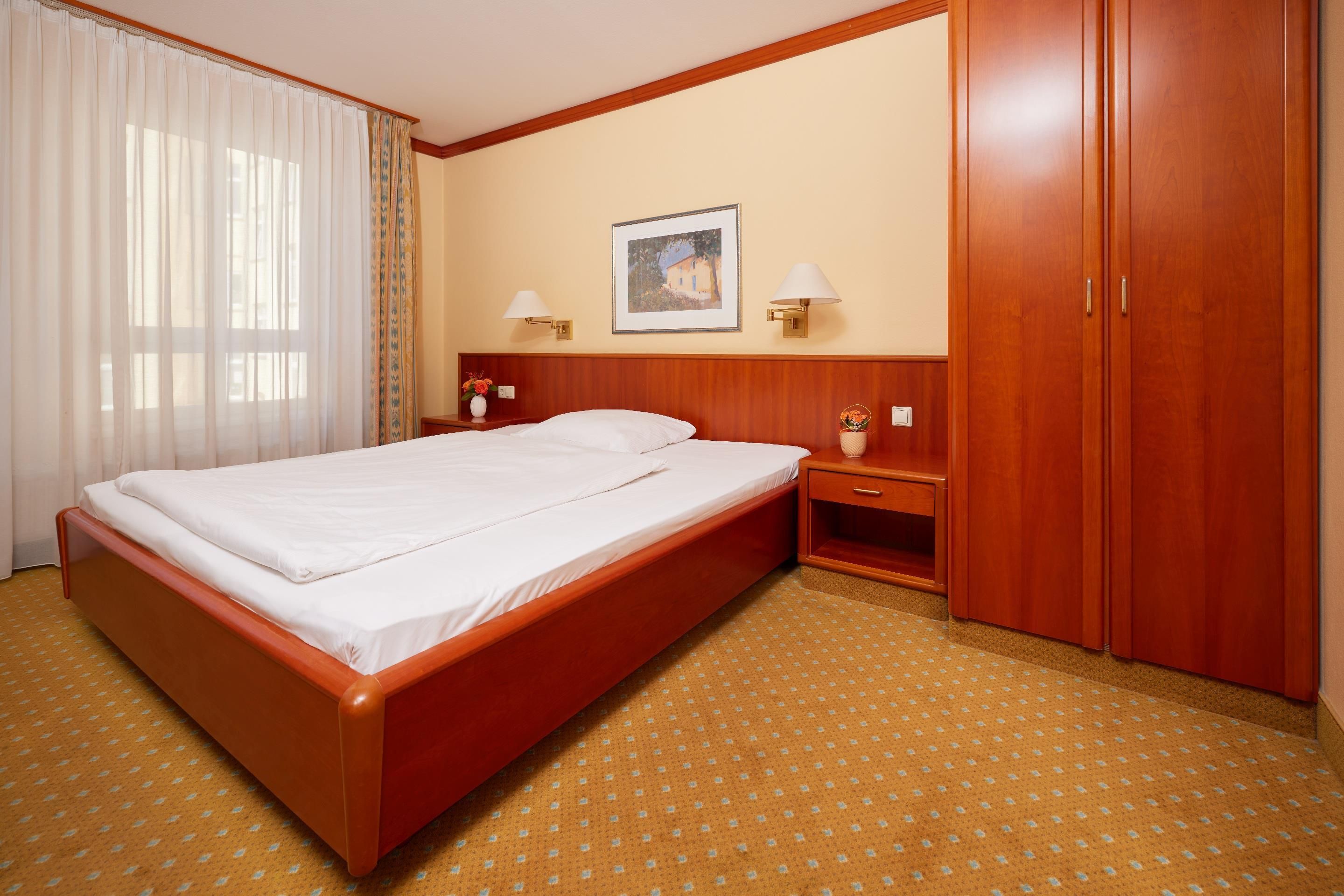 A hotel room with a large bed, wooden wardrobe, bedside tables with lamps, and a framed picture on the wall.
