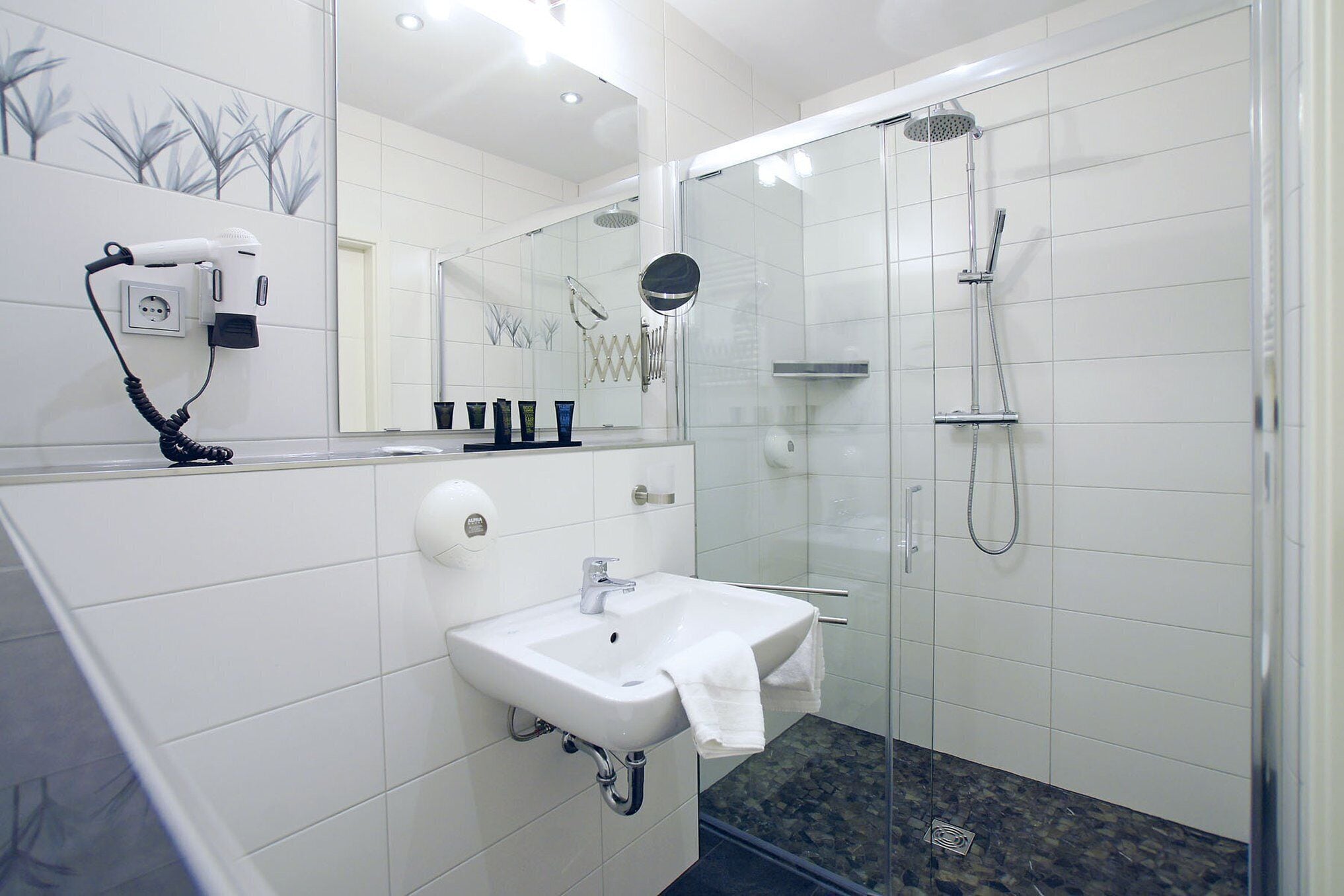 A modern bathroom with a glass-enclosed shower, a white sink, and a hairdryer mounted on the wall.