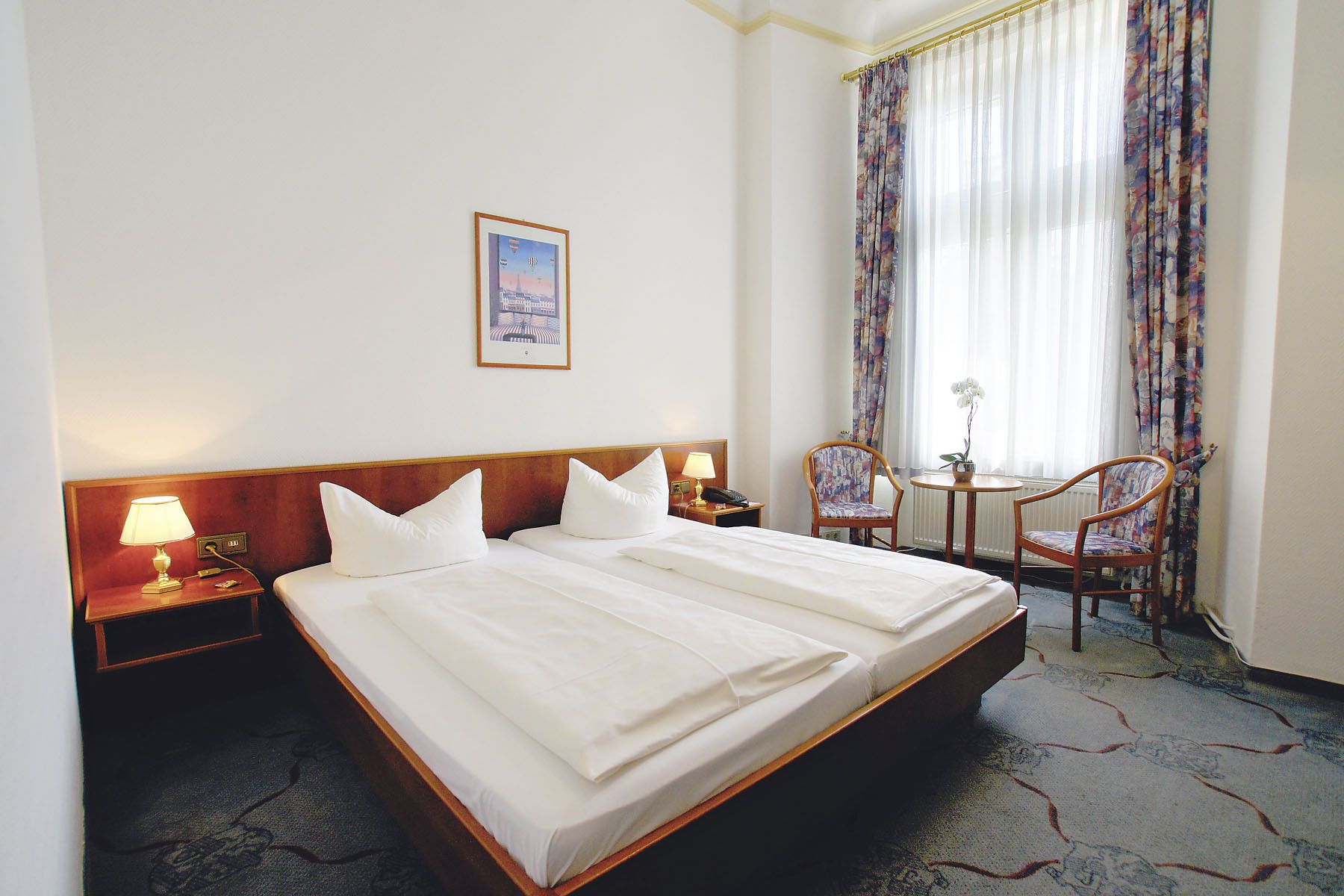 A hotel room with a large bed, two bedside tables with lamps, a chair, a small table with a flower, and a window with curtains.
