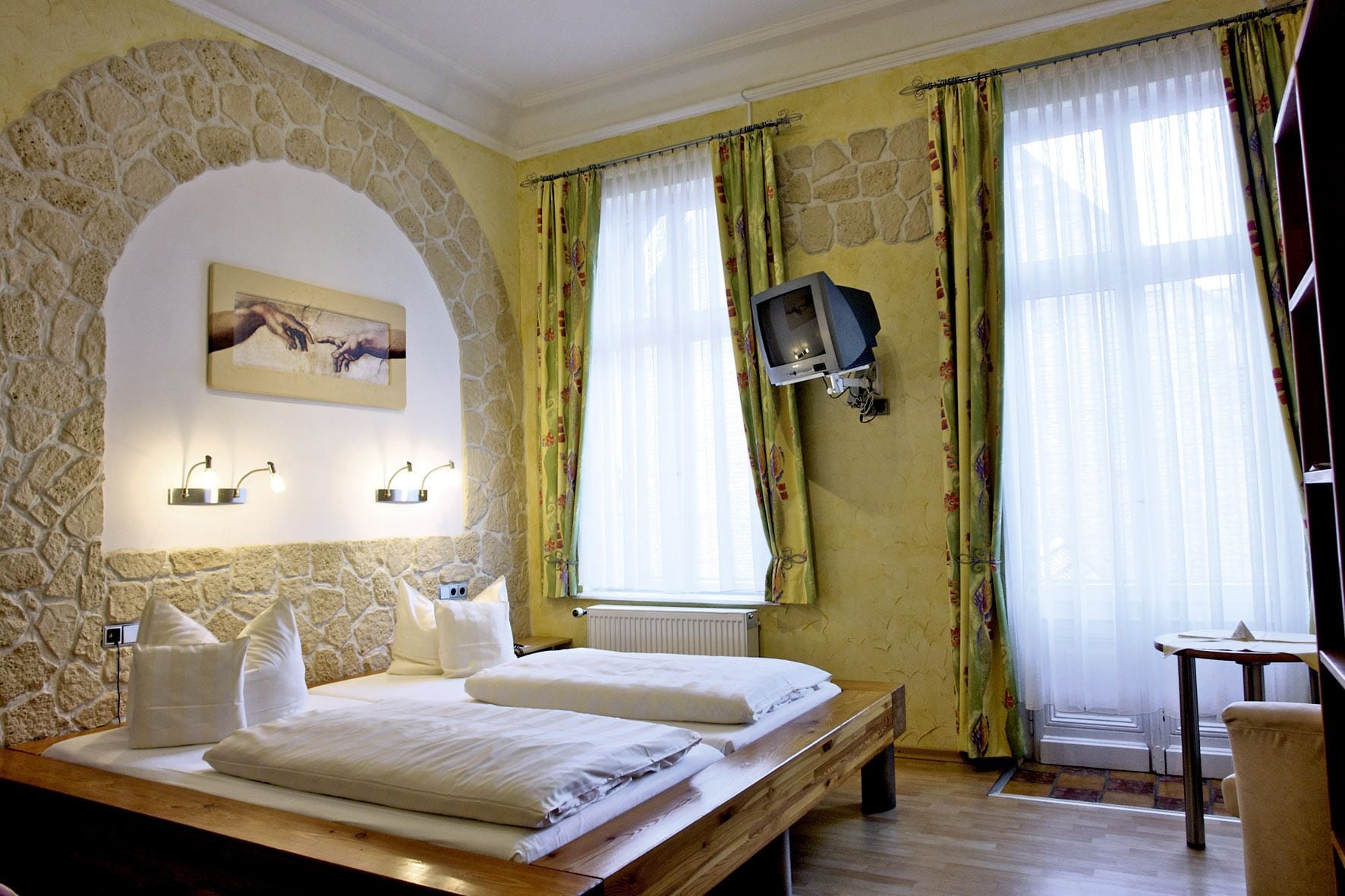A bedroom with a stone wall, a bed with white linens, a television mounted on the wall, and a window with curtains.
