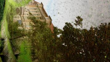 A historic building with a stone facade, surrounded by lush greenery and a body of water reflecting the structure.