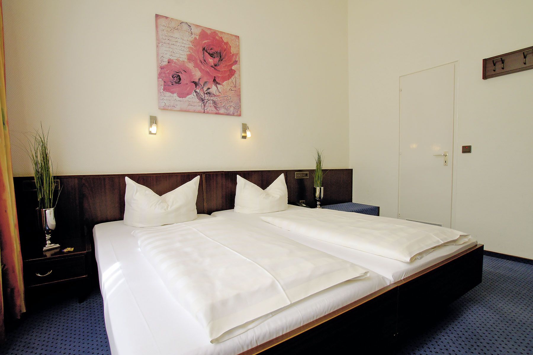 A double bed with white linens, a wooden headboard, a wall-mounted light, and a floral artwork above it.