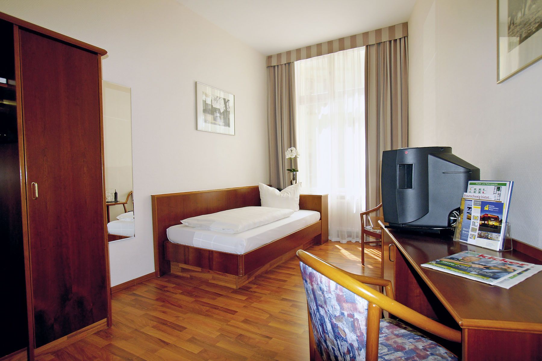 Habitación de hotel con cama de madera, un escritorio con televisor, una silla y una ventana con cortinas.