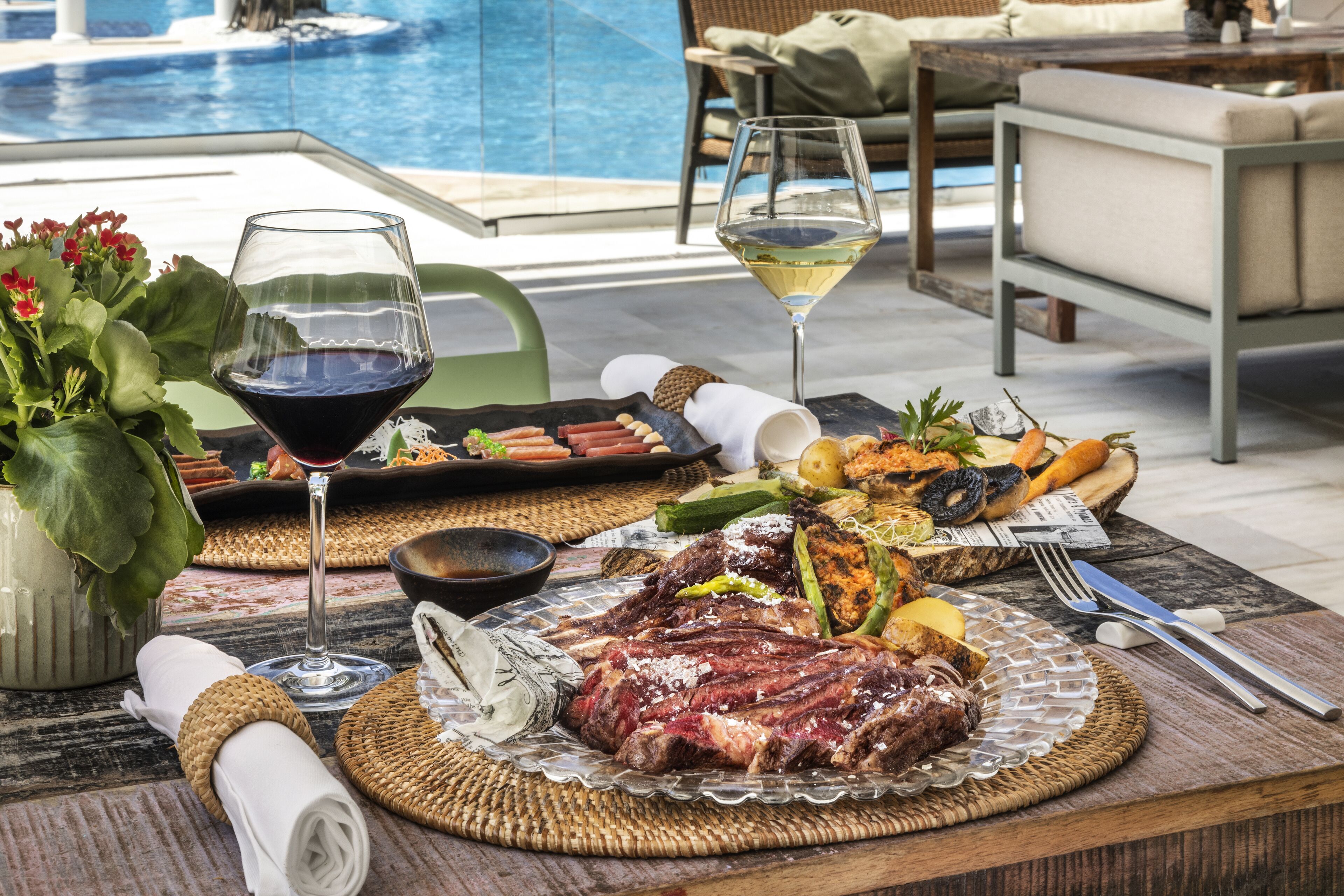 A dining table with a steak, vegetables, and wine glasses, set on a terrace with a pool view.