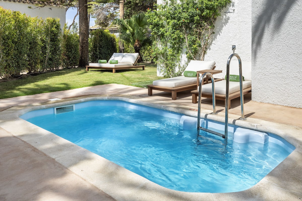 Ein rechteckiger Pool mit eingebauter Leiter, umgeben von einer Rasenfläche und einer weißen Mauer. Zwei Liegestühle mit Kissen und ein kleiner Tisch sind vorhanden.
