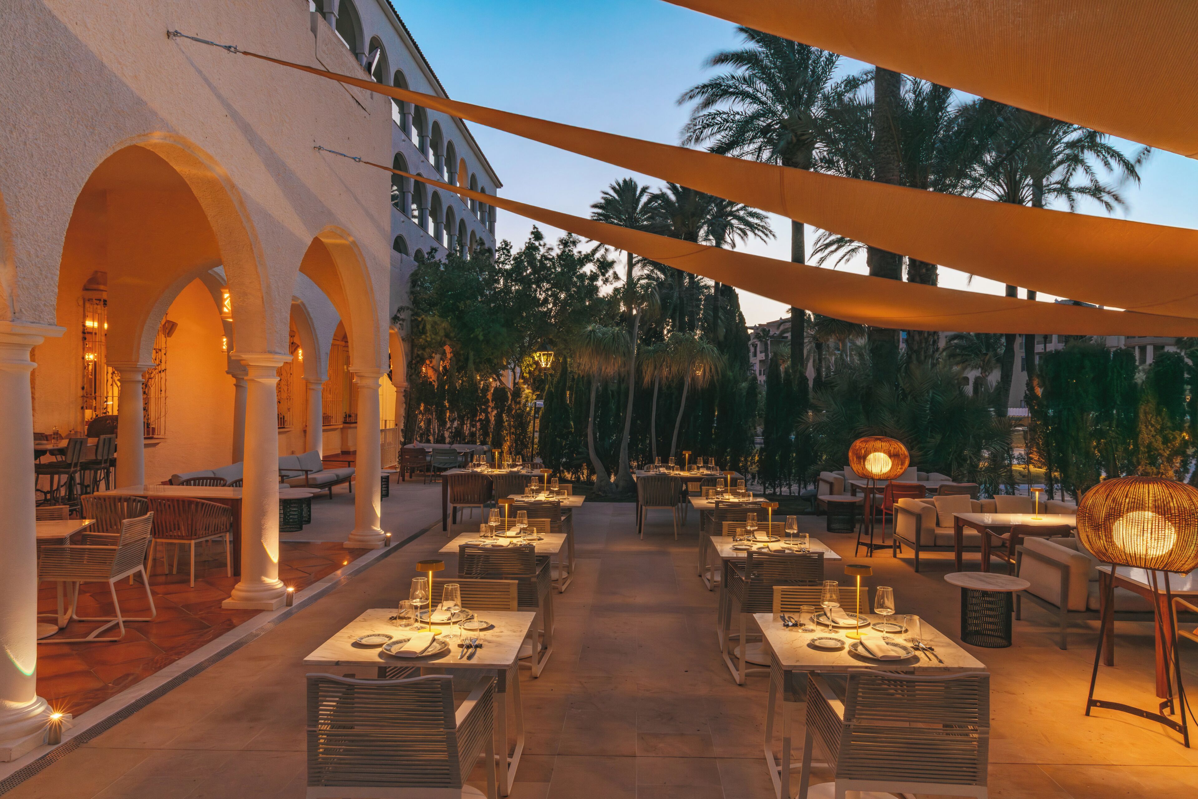 An outdoor dining area with tables and chairs, palm trees, and a building with arches.