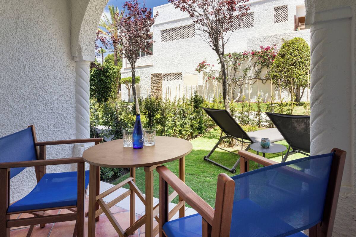 Eine Terrasse mit Holzkubeln und Tisch, umgeben von weißen Wänden und viel Grün.