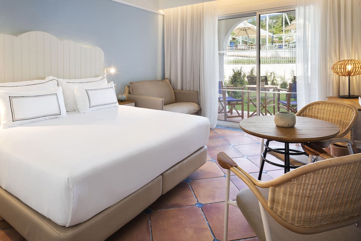 Ein Hotelzimmer mit einem großen Bett, einem Sessel, einem kleinen Tisch und Blick ins Freie.