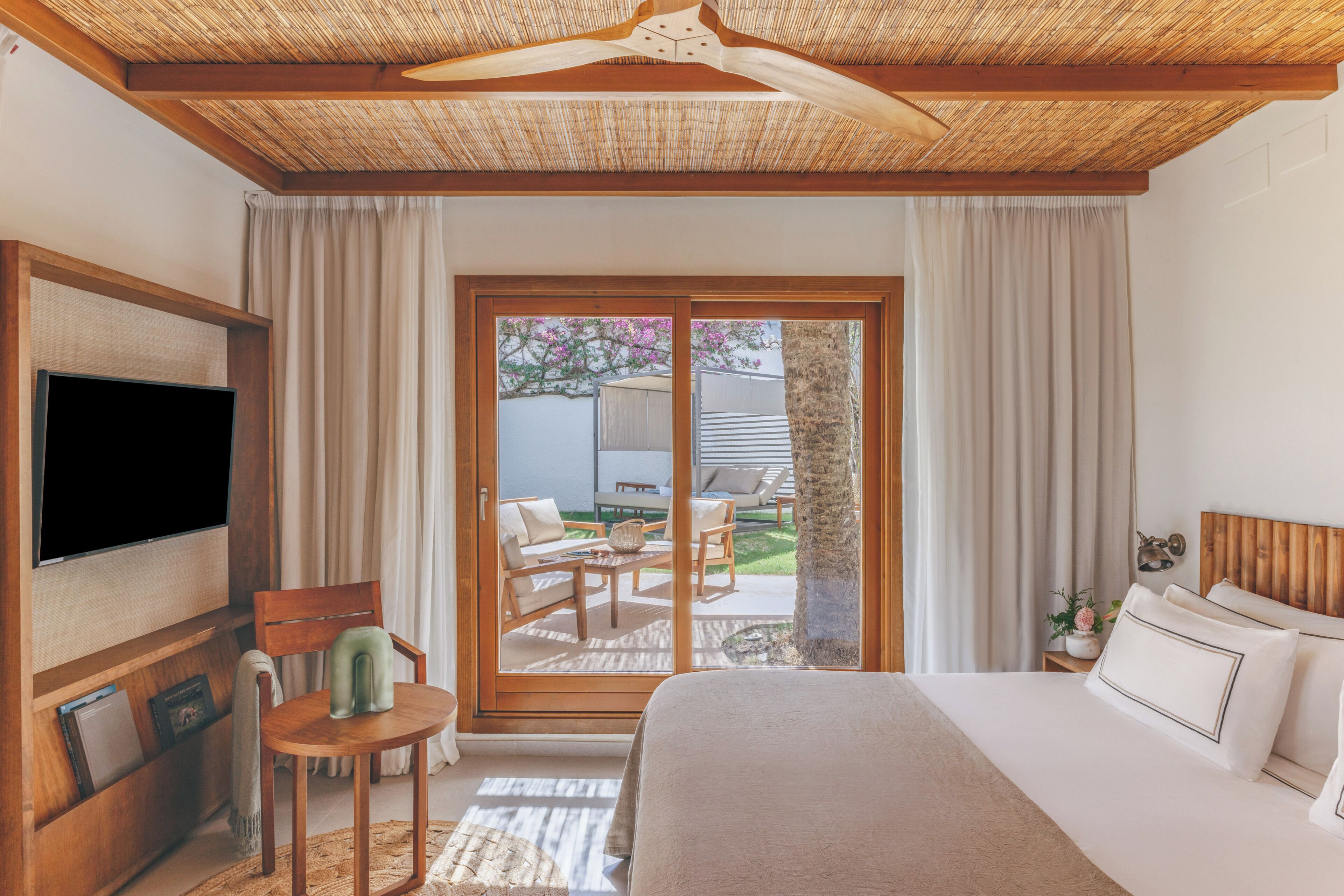 A bedroom with a bed, a TV, a chair, a small table, and a sliding glass door leading to an outdoor area with a table and chairs.