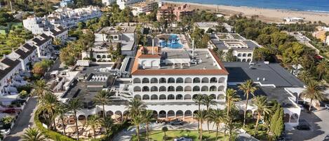 An aerial view of a resort with multiple buildings, a swimming pool, and a beach in the background.