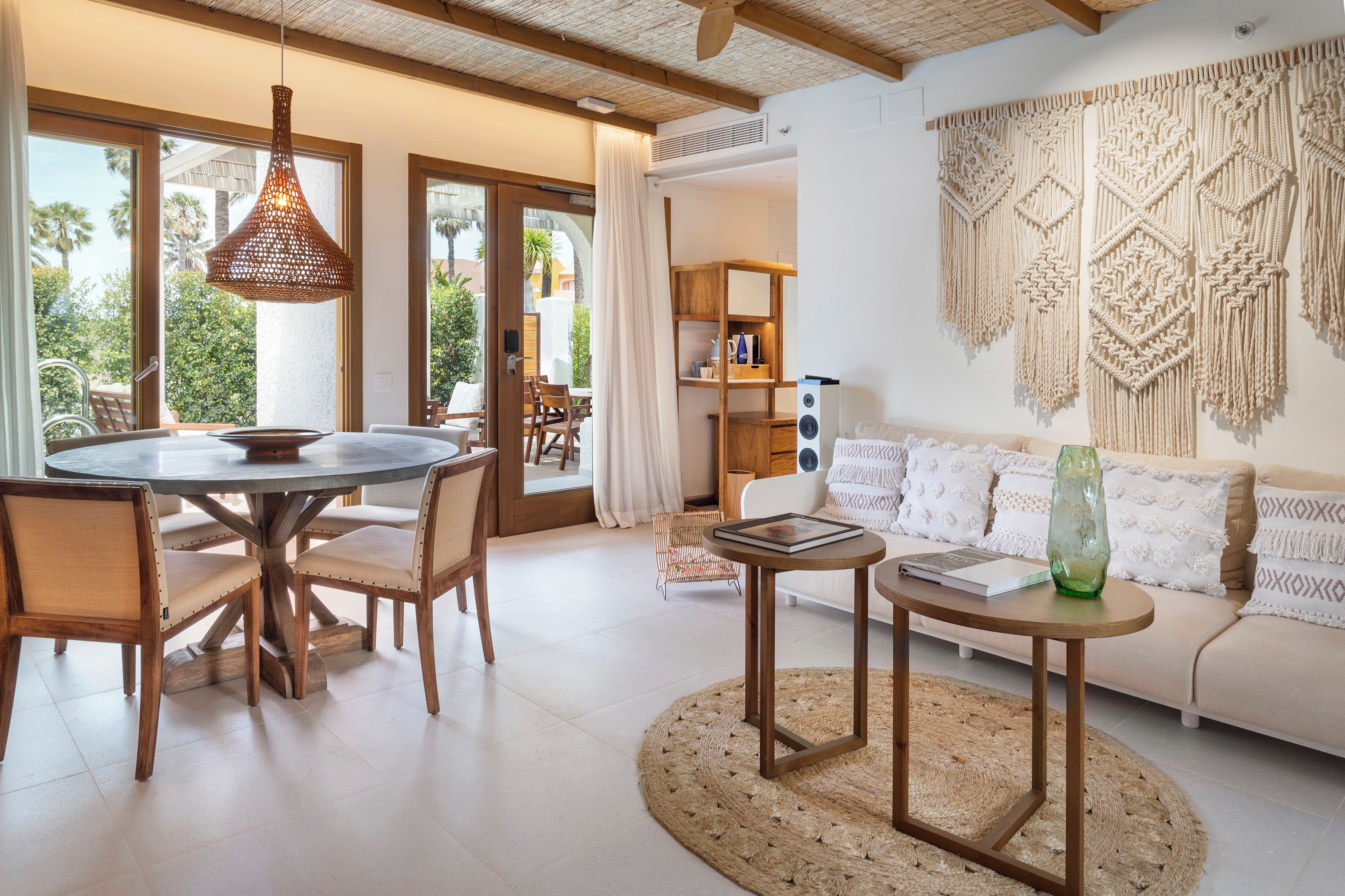 Uma sala de estar moderna com uma mesa de jantar redonda, cadeiras de madeira, um sofá e um painel de macramé na parede.