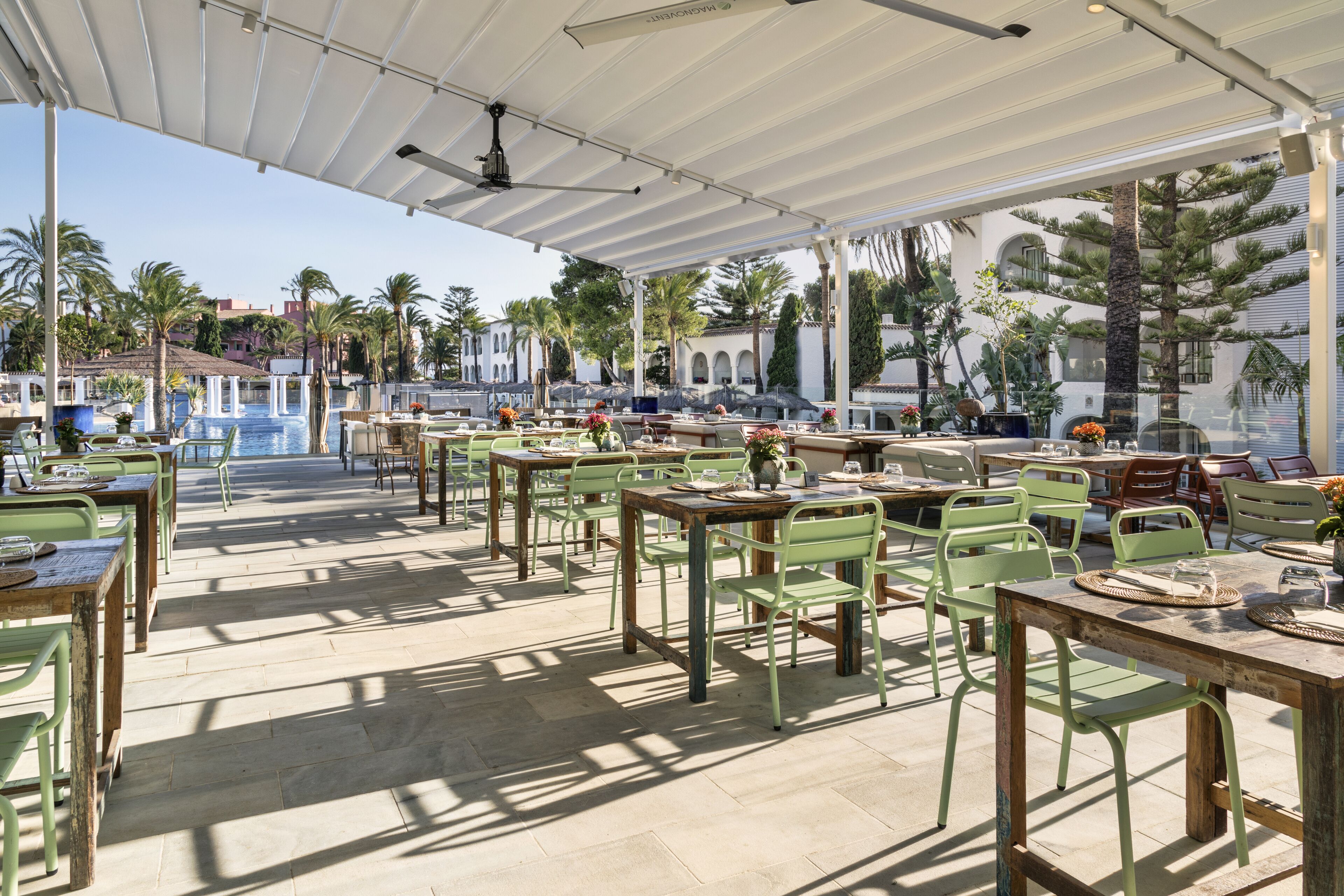Un área de comedor al aire libre con mesas y sillas, una piscina al fondo y palmeras.